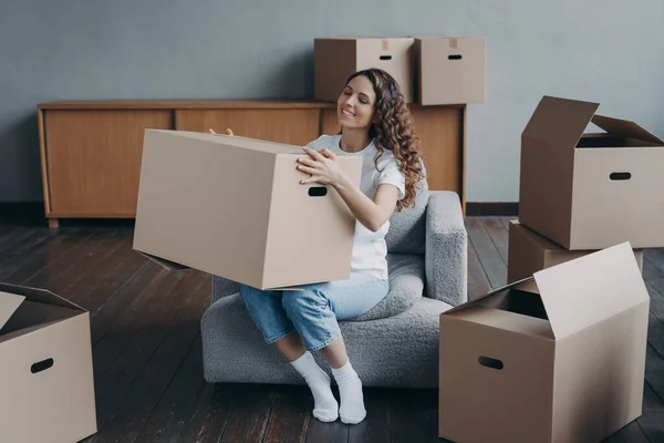 Proud european girl relocates to new home. Happy young woman moves alone. Lady is sitting in armchair among packed cardboard boxes. Independence, life choice and success concept.