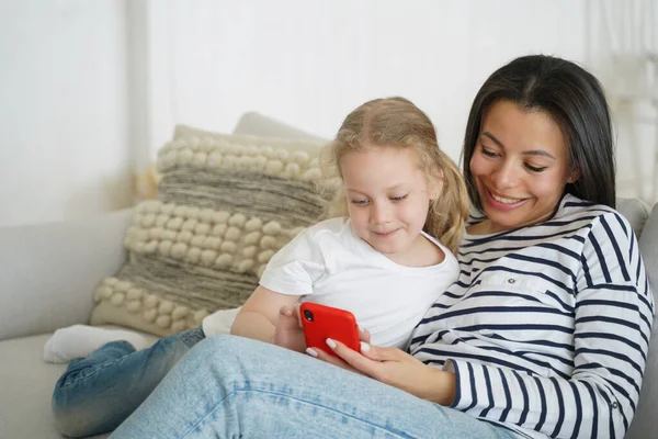 Mom and little daughter watching cartoons on smartphone, playing online game, sitting on couch. Mother and small girl use learning mobile app enjoying leisure time on sofa together.