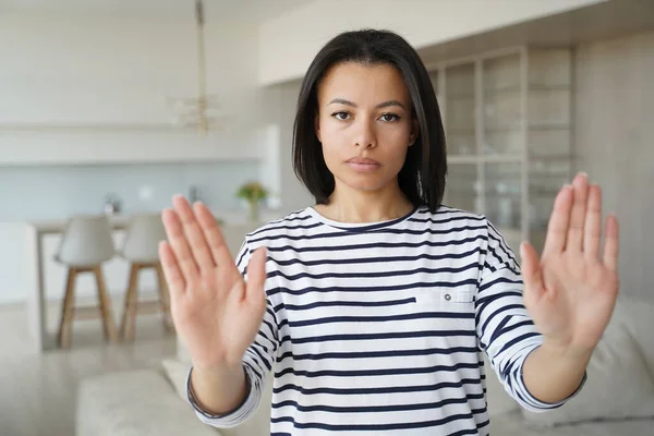 Serious young woman showing stop hand warning gesture, protesting against domestic violence or abortion. Strict female says no to abuse and discrimination, expressing disagreement, standing at home.