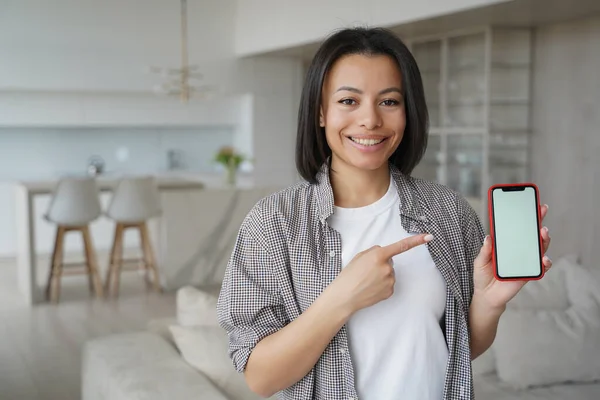 Smiling female tenant pointing at smartphone with mockup blank screen at home. Happy woman renter showing template phone screen, recommends house hunting app, service for housing search.
