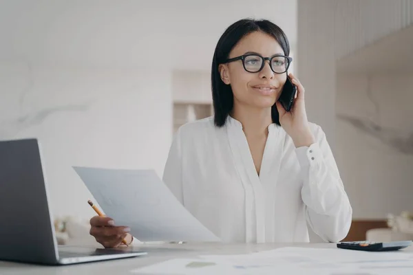 Young hispanic businesswoman in glasses has phone talk. Girl is working remote at home office on quarantine. Manager, accountant or business assistant is doing paperwork using laptop, examining data.