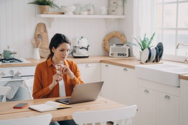 Mutfak masasında oturan meşgul bir kadın, evde dizüstü bilgisayarda çalışıyor. Düşünceli kadın internetten alışveriş yapıyor, alışverişleri planlıyor, yapılacaklar listesi yapıyor, internetten dil öğreniyor. Uzak eğitim, uzak iş.
