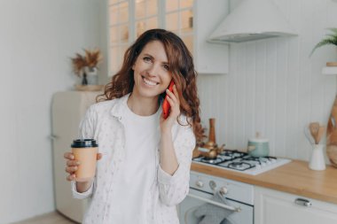 Gülümseyen genç bir bayan elinde bir fincan paket kahve ile evdeki rahat modern mutfaktan hoş bir cep telefonu görüşmesi yapıyor. Akıllı telefondan konuşan, iletişimin tadını çıkaran mutlu bir kadın. Verimli bir gün.