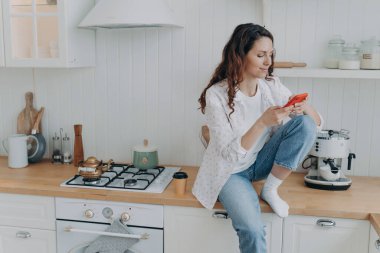 Akıllı telefon tutan, sosyal ağlarda sohbet eden, modern, rahat bir mutfakta oturan bir kadın. Kadın angarya işlerden sonra telefonuyla dinleniyor, internetten alışveriş yapıyor. E-ticaret, ev hayatı konsepti.