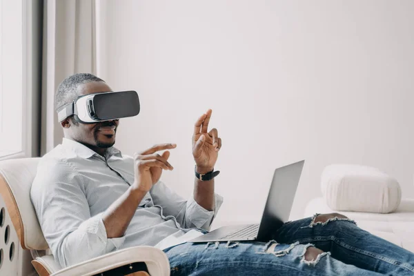 African american male software developer in virtual reality glasses tests new app, working on laptop, sitting in chair. Modern businessman touching objects in cyberspace, uses high tech for business.