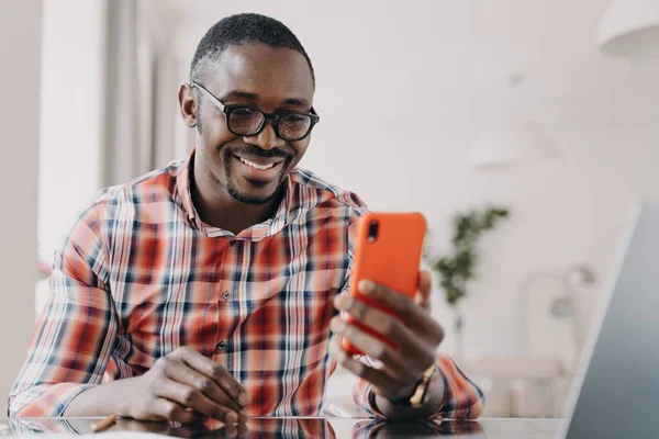 Modern african american man wearing glasses uses mobile apps, holding smartphone, sitting at desk smiling. Happy young black guy looking at phone, reading good news message or watching funny video.