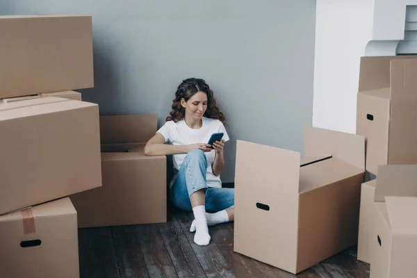 Happy young european woman with smartphone is packing boxes. Girl is chatting on mobile phone and sitting on the floor. Easy moving and shipping service order concept.