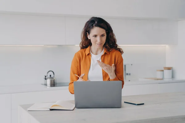 Hispanic female teacher in earbud working at laptop, conducts language lesson by video call, sitting in kitchen at home. Businesswoman chatting with colleague or client online on computer. Remote job