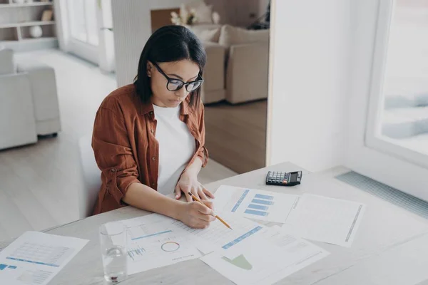 Top down view of businesswoman examining reports a the desk at home. Young hispanic woman in glasses is working at home office on quarantine. Business assistant, freelancer is working on project.