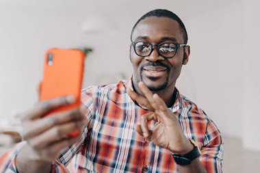 Gülümseyen Afro-Amerikalı adam onaylayıcı bir jesti eliyle tutuyor, akıllı telefon tutuyor, video aracılığıyla iletişim kuruyor. Modern siyah adam gözlük takıyor, telefon ekranına bakıyor, tamam işareti gösteriyor..