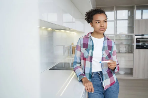 Pensive teenage girl gets positive pregnancy test result. Sad african american young woman is holding stripe test. Teenage lifestyle, unwanted pregnancy and motherhood. Contraception concept.
