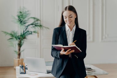 Siyah takım elbiseli, güler yüzlü genç iş kadını elinde not defteri ve yazı puanları, ofiste dizüstü bilgisayarla beyaz masaya yaslanmış, arka planda palmiye ağacı var.
