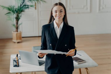 Siyah takım elbiseli, güler yüzlü genç bir kadının portresi. Beyaz masanın önünde dizüstü bilgisayarı açık ve arka planında palmiye ağacı olan bir kadın..