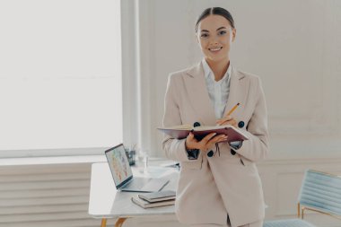 Zarif ve mutlu bir kadın yönetmen, planlama çalışma gününde, modern ışık ofisinin içinde mutlu bir şekilde dururken, evde uzaktan çalışırken notlar alan mutlu bir iş kadını.