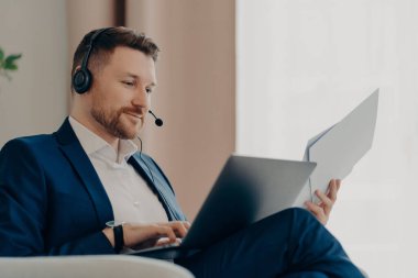 İş adamıyım. Sırıtkan sakallı erkek patronun kucağında evrak ve dizüstü bilgisayarla evde oturmuş online toplantı yapıyor. İş adamları ve modern teknolojiler