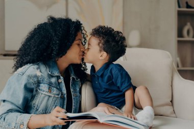 Güzel Afro-Amerikan aile annesi ve küçük oğlu birbirlerini öpüyor ve birlikte evde vakit geçirirken sevgisini ifade ediyor oturma odasındaki yumuşak beyaz koltukta oturup kitap okuyor.
