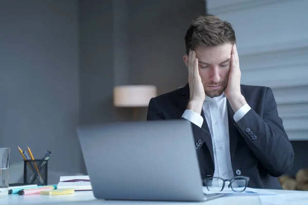 Exhausted man German legal consultant sits at workplace at home office in formal suit, took off glasses, gently touches his temples with fingers, feels headache while working in online court session