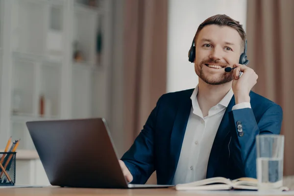 Young positive businessman in suit sitting at the table at home office wearing headset while organizing online conference on laptop, male freelancer having video call with team. Remote work concept