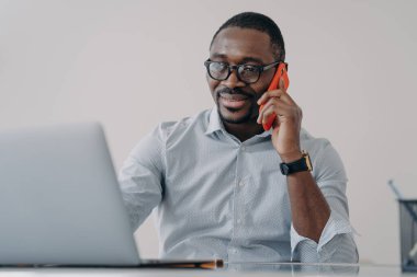 Afrikalı Amerikalı iş adamı iş görüşmesi yapar, telefonla konuşur, dizüstü bilgisayarda çalışmayı tartışır. Gözlüklü, gülümseyen siyah erkek yönetici. Akıllı telefon, danışman müvekkil, ofis masasında oturuyor..