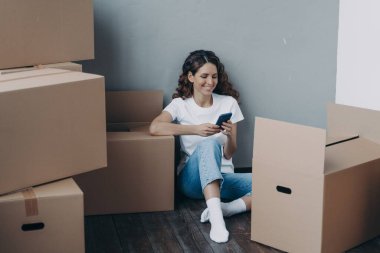 Mutlu Latin kadın cep telefonu uygulamaları kullanıyor, taşınma servisini seçiyor, taşınmaya hazırlanıyor, nakliyecisine çevrimiçi cevap veriyor, karton kutularda paketlerle yerde oturuyor. Kolay taşınma günü