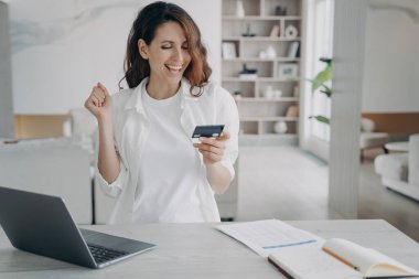 Banka kartı tutan heyecanlı kadın dizüstü bilgisayarında online bankacılık hizmeti kullanıyor. Mutlu kadın evet hareketi yapar, nakitten memnun olur, kolay ödeme yapılır. E-bankacılık, e-ticaret.