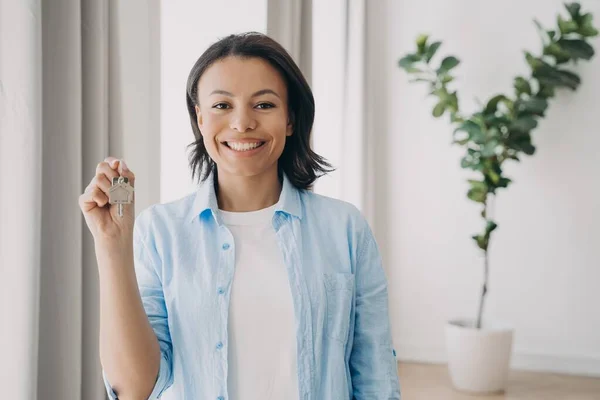 Gorgeous young lady is buying apartment and moving. Satisfied mixed race woman is holding house keychain in her hand near window of living room. Mortgage loan and investment concept.
