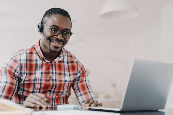 Smiling african american man in wireless headset sits at desk, studying online on laptop, happy modern young black guy in headphones watching webinar on computer. E-learning, distance education.
