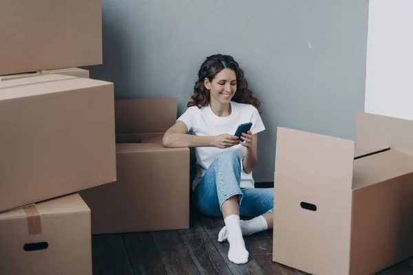 Happy latina female uses mobile phone apps, choosing moving service, preparing for relocation, answer her mover online, sitting on the floor with packed things in cardboard boxes. Easy moving day