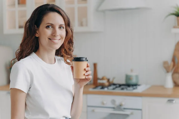 Portrait of friendly happy hispanic woman holding disposable paper cup of coffee in modern kitchen. Smiling relaxed female housewife taking a break during chores, enjoying hot drink at home.