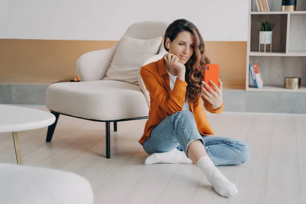 Modern woman, holding smartphone, taking selfie photo or chatting by video call, sitting on heated floor in living room. Female wearing earphones communicates online using mobile phone app at home.