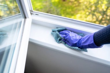 Temizlik ve temizlik konsepti. Mor eldivenli genç bir kız pencere pervazındaki tozu bir bezle siliyor.