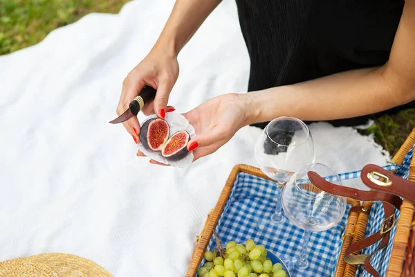 Romantic picnic in the park on the grass, delicious food: basket, wine, grapes, figs, cheese, blue checkered tablecloth, two glasses of wine. Girl cuts figs.Outdoor recreation concept