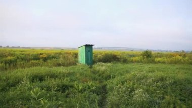 Old wooden toilet with a carved window with the shape of a heart cut out on the door, in open field. Vintage WC. An outdoor rustic green restroom in a field landscape of flowers and grass at sunrise
