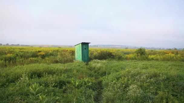 Starý dřevěný záchod s vyřezávaným oknem ve tvaru srdce vyříznutého na dveřích, na otevřeném poli. Ročník WC. Venkovní venkovské zelené toalety na poli krajiny květin a trávy při východu slunce — Stock video