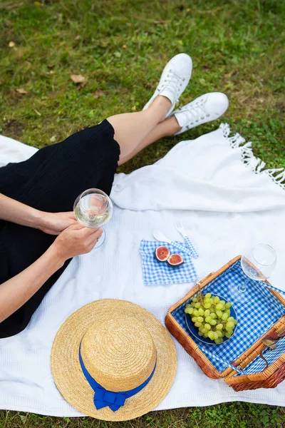 Romantic picnic in the park on the grass, delicious food: basket, wine, grapes, figs, cheese, blue checkered tablecloth, two glasses of wine. The girl is holding a glass of wine in her hands.The concept of outdoor recreation