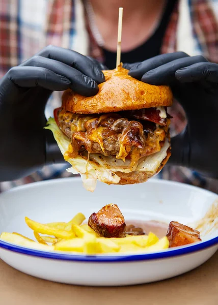 The concept of fast food and takeaway food. A man in black latex gloves holds a juicy hamburger in his hands, lies near french fries on a metal plate along with cheese sauce. Close-up