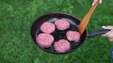 Meat for hamburgers grilling on background of grass. Fry the cutlets in a pan with oil. Woman hand flips a meatballs for burger with a spatula - barbecue during beautiful summer afternoon outdoors