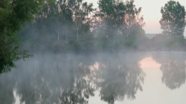 Sunrise over lake and beautiful morning landscape. Early morning sun breaks through the thick fog in summer forest near the rural reservoir. Fantastic foggy river in golden sunlight on water surface