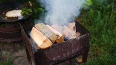 Firewood is burning in the grill in outdoor. Kindling firewood on a summer evening for barbecue. Wood logs are smoking in the metal brazier close up. White thick smoke rises from the roaster