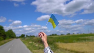 Ukrainian flag is fluttering on blue sky background with clouds, road and wheat field. Girl holding national flag in her hand - celebrating Independence Day. Pray for Ukraine and stop war concept