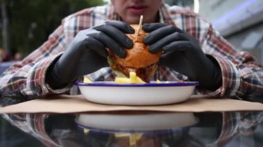 Ekose gömlekli ve siyah eldivenli bir adam sulu bir burgerden ısırıp çiğniyor. Sos, adamın bir tabak patates kızartmasının üzerinde tuttuğu hamburgerden damlıyor. Açık bir masada oturuyor. Yakın çekim.