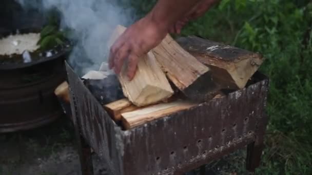 木头原木在金属钎子里冒着烟 一个男人在自家后院把柴火扔进了一个冒烟的烤架里 烤面包机里升起浓浓的白烟 篝火在户外的烤架上燃烧着 — 图库视频影像