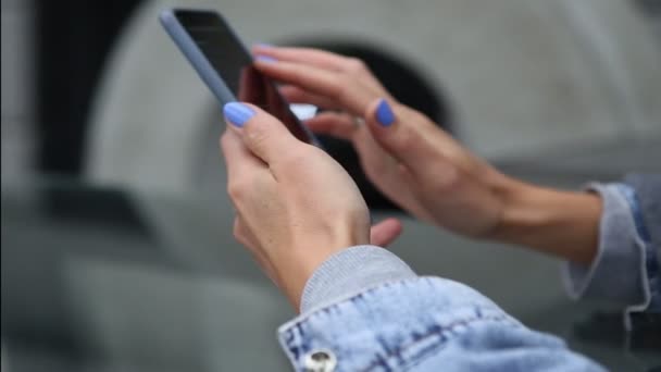 携帯電話の青いマニキュアタッチスクロールページアプリを持つ女性の指 女の子でデニムジャケット検索や屋外で映画をオンラインで見るためにインターネットライブラリ内のムービービデオを閲覧 — ストック動画