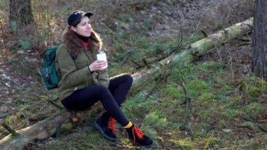 Beautiful girl with a backpack drinks tea from a thermos in the forest sitting on a log. Woman warming up by drinking hot drink. Female traveler sitting and relaxing in nature in spring among trees