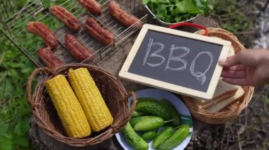 Bir kadın eli, ızgarada sosis, sepette mısır, salatalık ve roka olan ahşap bir kütüğün üzerine barbekü tahtası koyar. Güneşli bahçede üst manzara.
