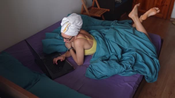 ベッドの上にノートパソコンに横になってキーボードを押す女性 頭にタオルでシャワーした後の笑顔の女の子は嘘をつき 彼女の足でノートブックで動作します 隔離室で働いてる — ストック動画