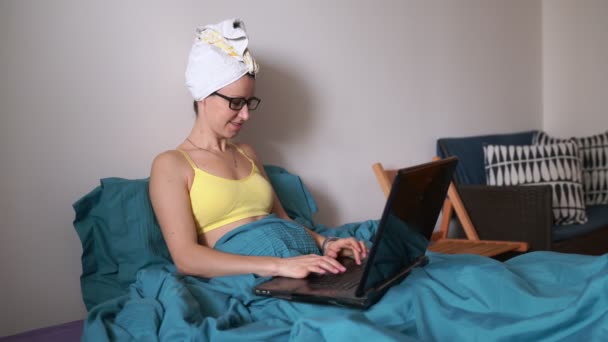 快乐美丽的年轻女人 头上拿着毛巾 坐在床上的笔记本电脑上工作 迷人的女孩 脸上挂着可爱的微笑 躺在床上 躺在笔记本上聊天 — 图库视频影像