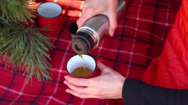 Kadın eli termostan sıcak bir içecek dolduruyor. Tema seyahati. Gezgin kız termos bardağından çay doldururken dışarıda ekoseli bir yerde oturuyor. Yürüyüş sırasında kırmızı fincanda çay içen genç bir kadın.