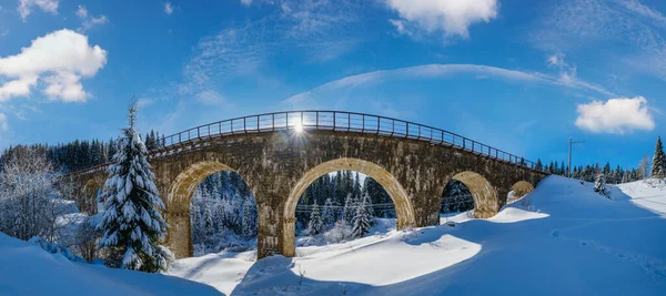 Stone viaduct (arch bridge) on railway through mountain snowy fir forest. Snow drifts  on wayside and hoarfrost on trees and electric line wires.