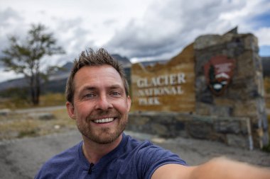 Gömleğine mikrofon takmış biri Buzul Ulusal Parkı 'nın doğu girişindeki tabelasının önünde selfie çekiyor.